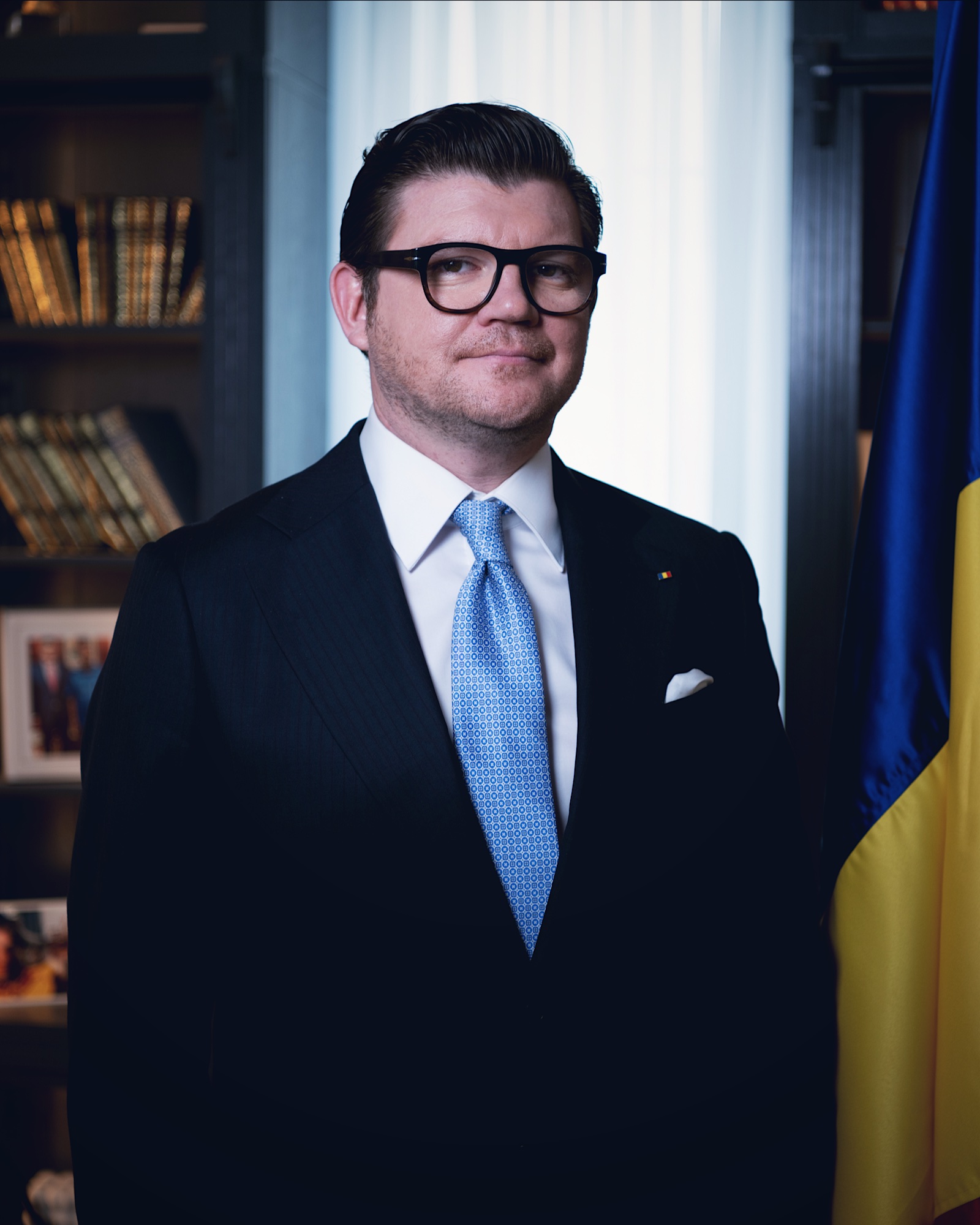 Anton Pisaroglu in a dark suit and blue tie, standing beside the Romanian flag.