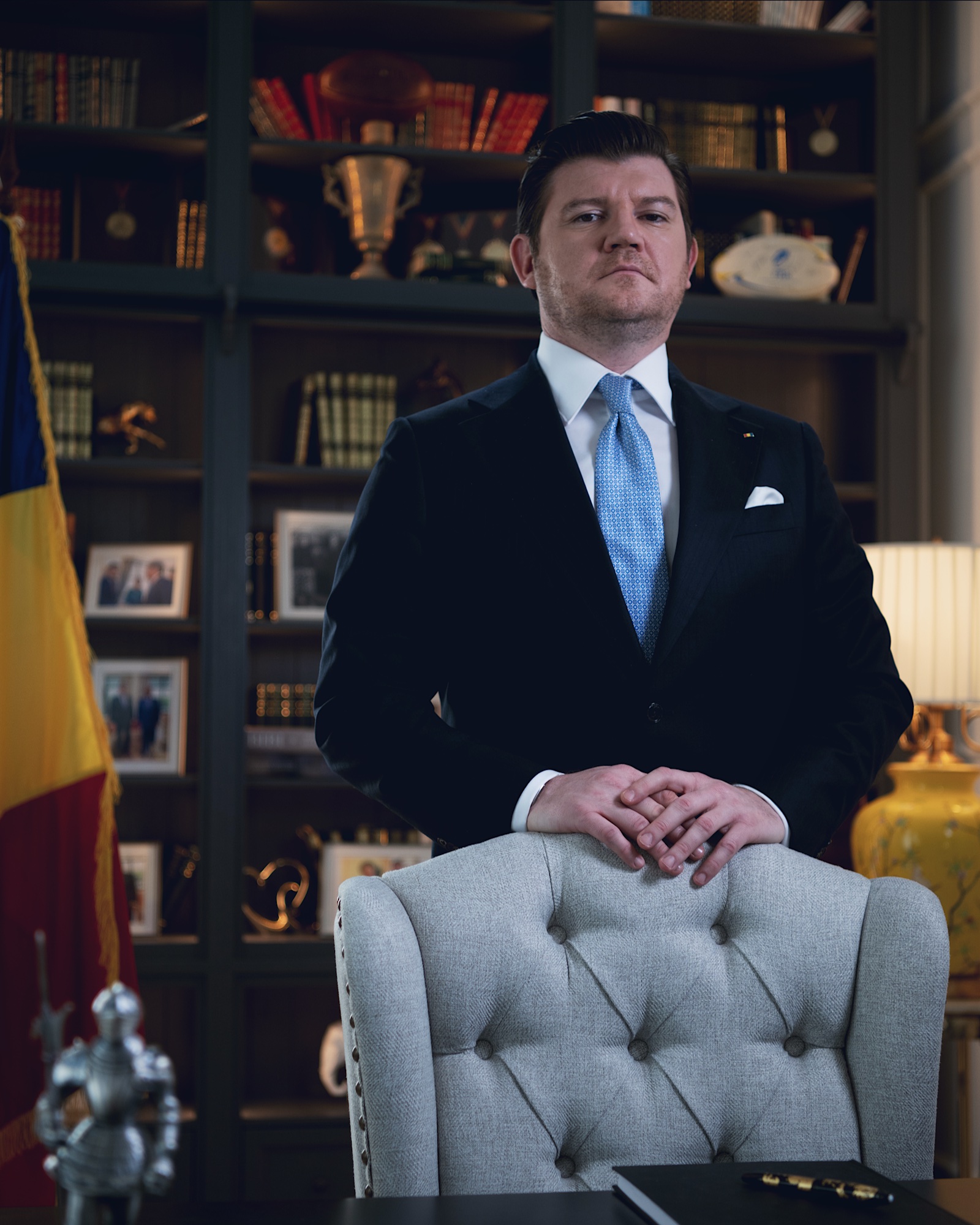 Anton Pisaroglu standing behind a grey upholstered chair in a dark suit and blue tie, with bookshelves behind him.