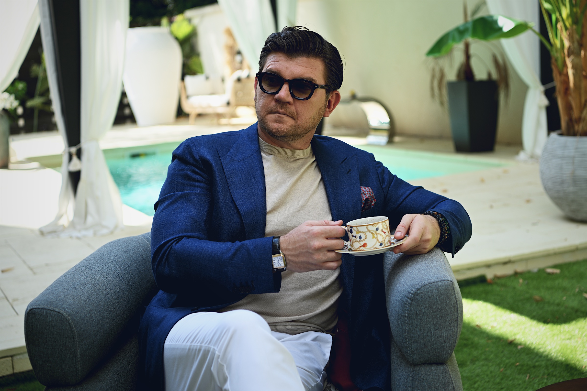 Anton Pisaroglu sitting beside a pool in a blue blazer and sunglasses, holding a patterned cup and saucer.
