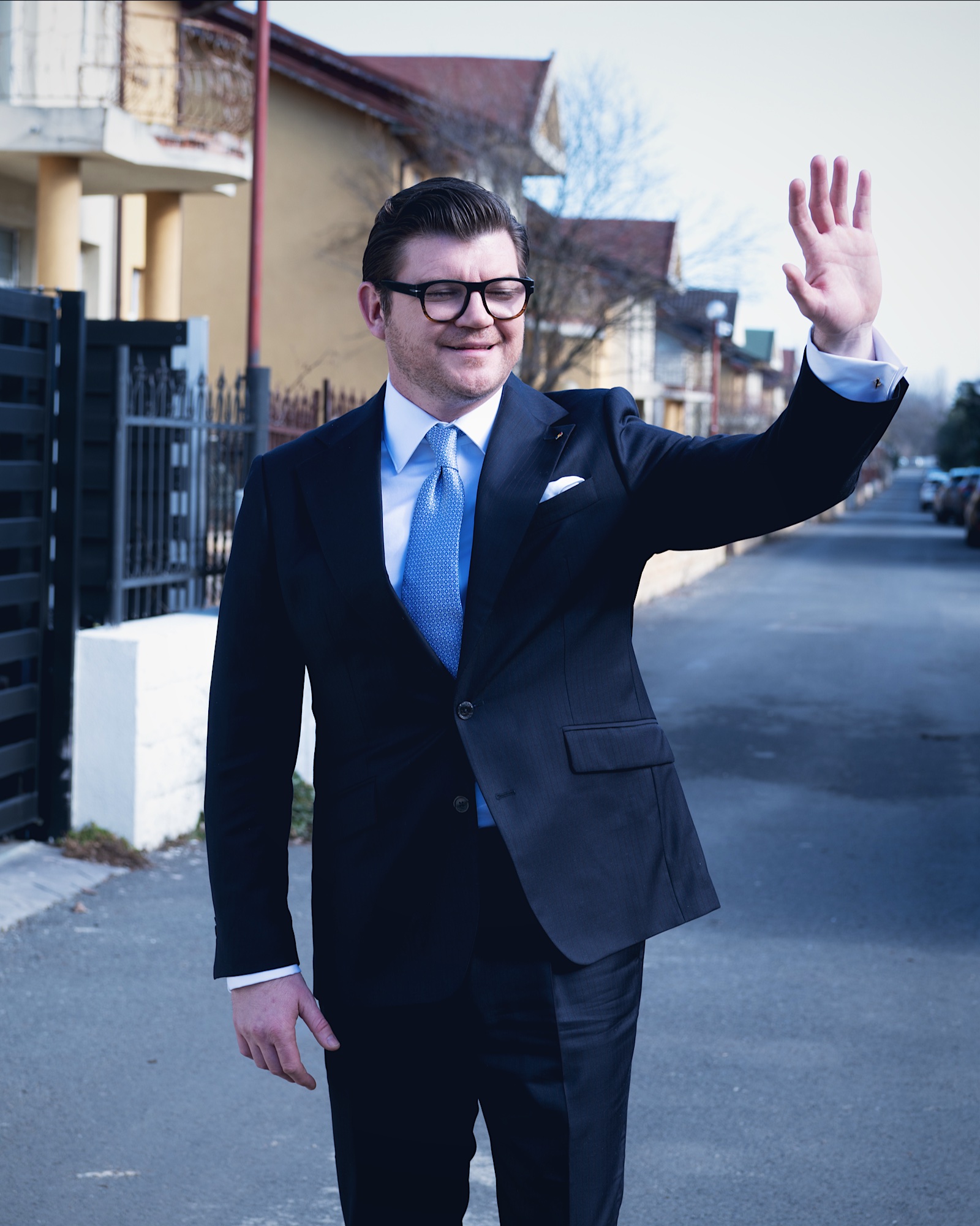 Anton Pisaroglu waving on a residential street in a dark suit and blue tie.