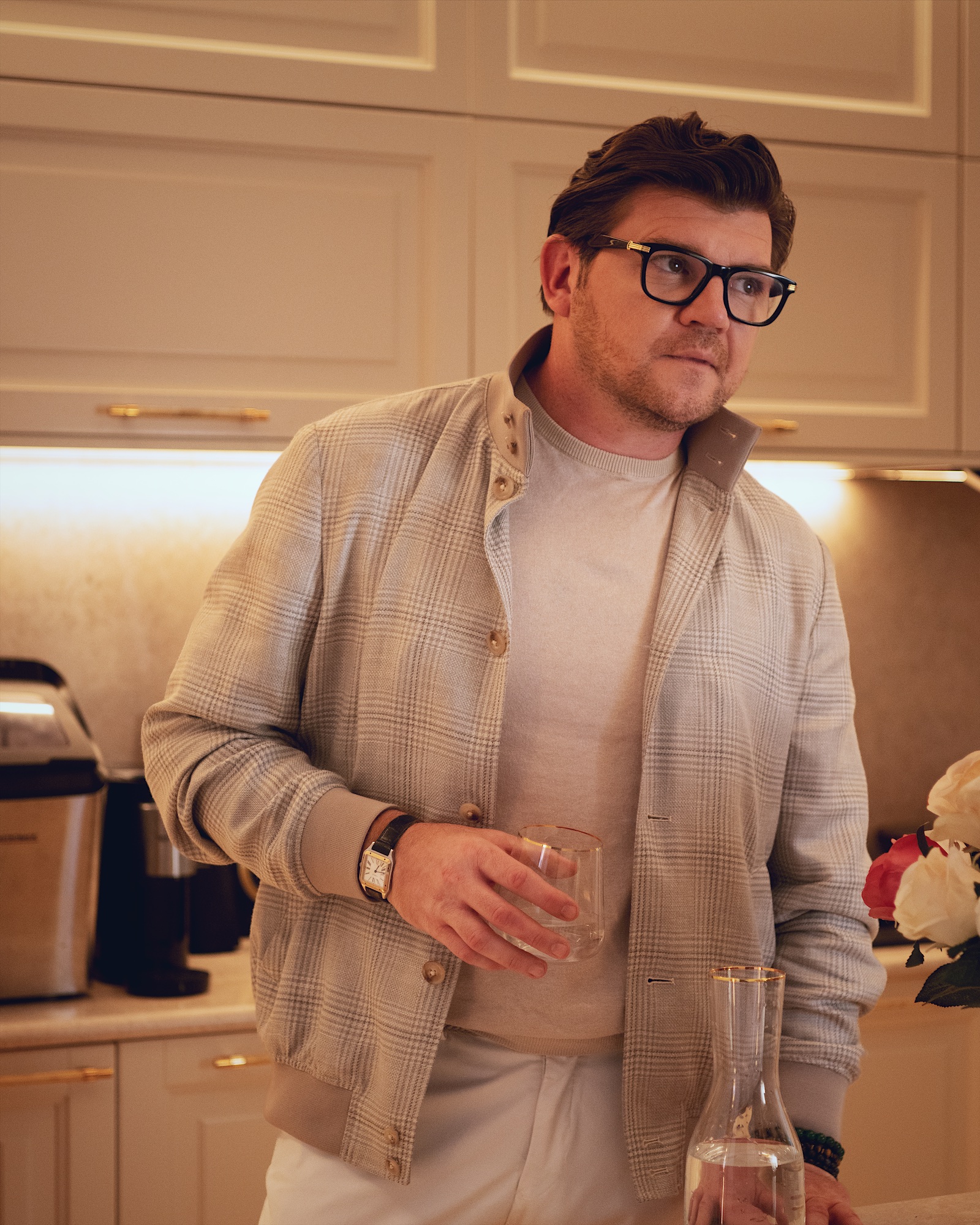 Anton Pisaroglu standing at a kitchen counter in a light checked jacket, holding a glass beside a carafe.