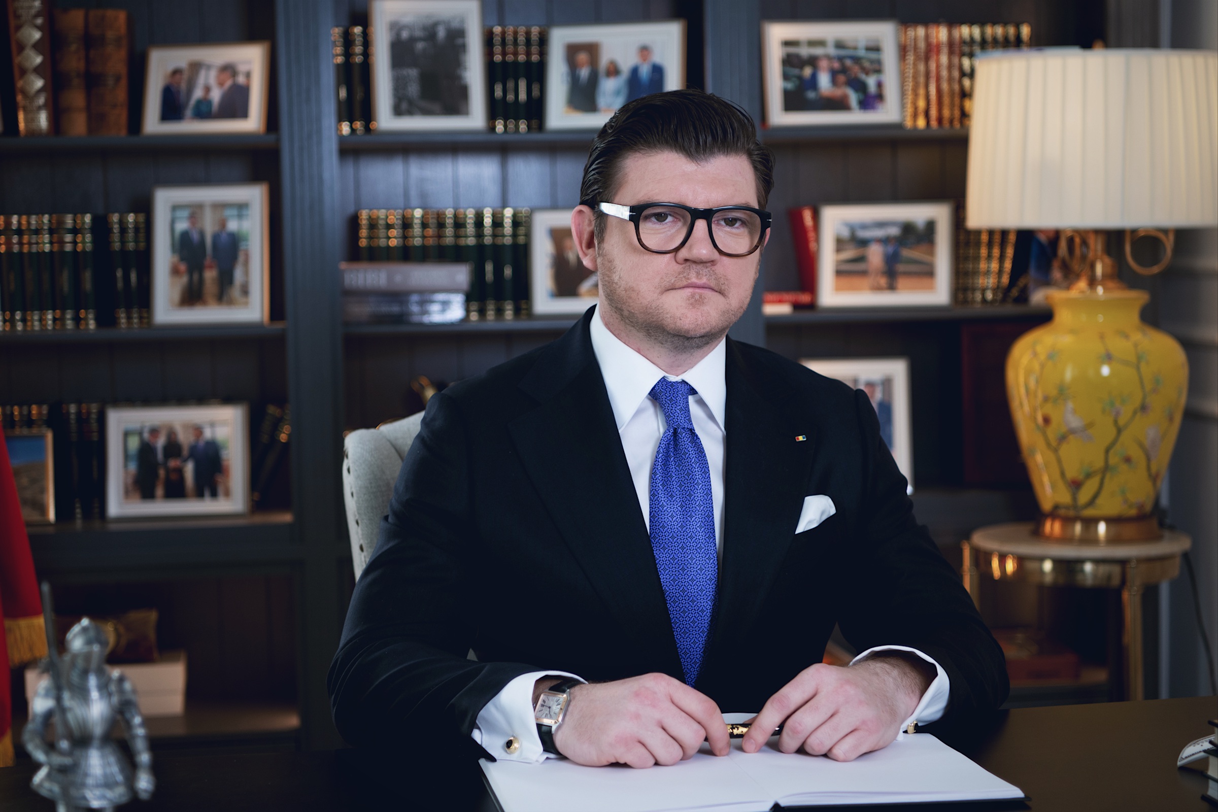 Anton Pisaroglu sitting at a desk with a pen and an open notebook, with bookshelves and framed photographs behind him.