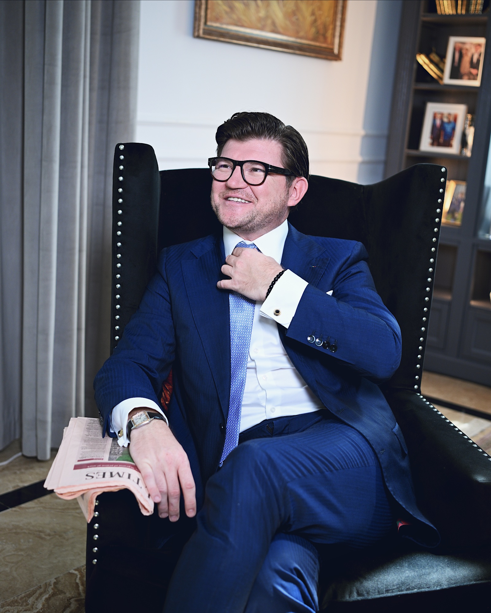 Anton Pisaroglu smiling in a dark wingback chair, with one hand at his tie and a folded newspaper on the armrest.