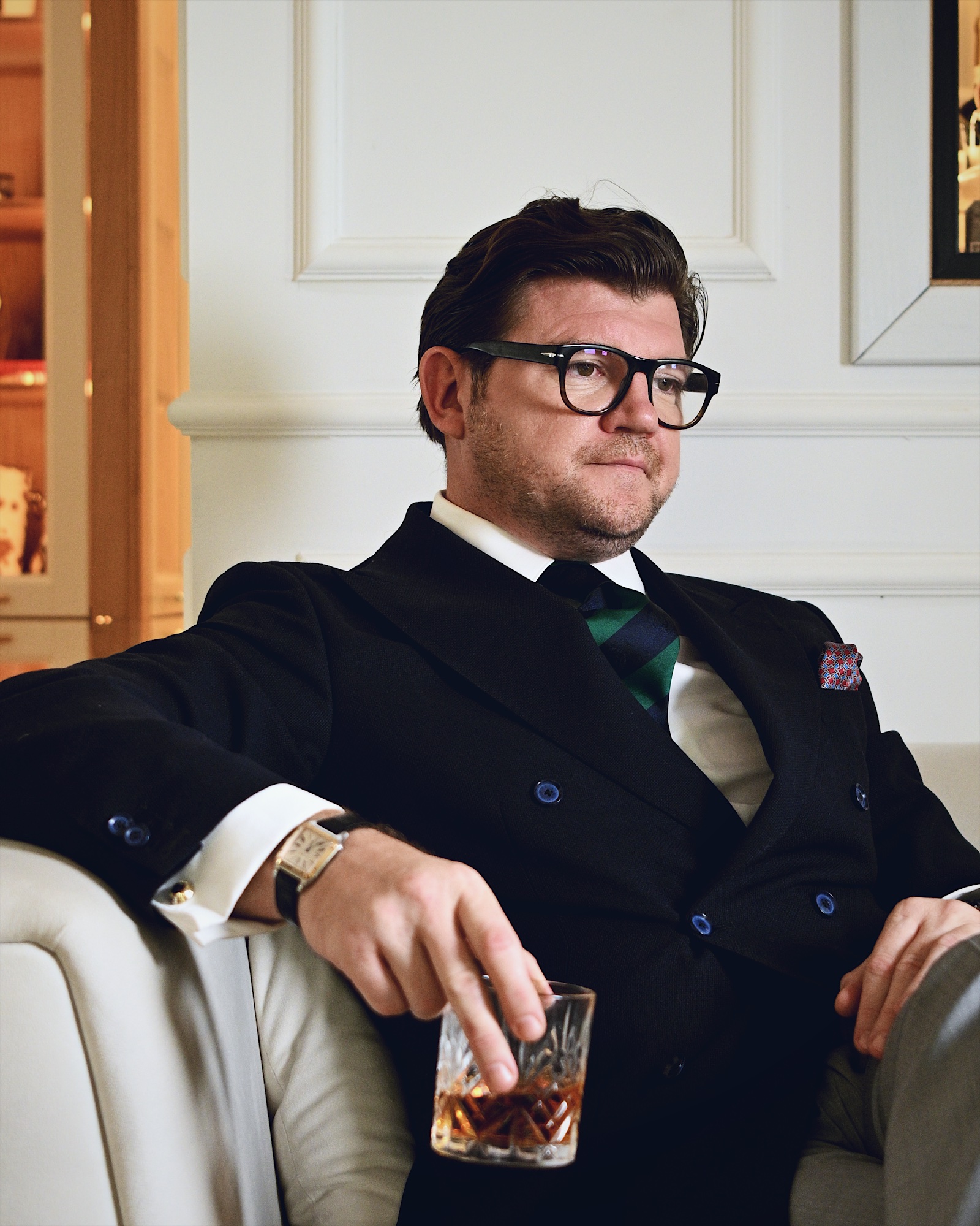 Anton Pisaroglu sitting in a cream armchair, wearing glasses and a dark double-breasted blazer, with a patterned drinking glass in one hand.