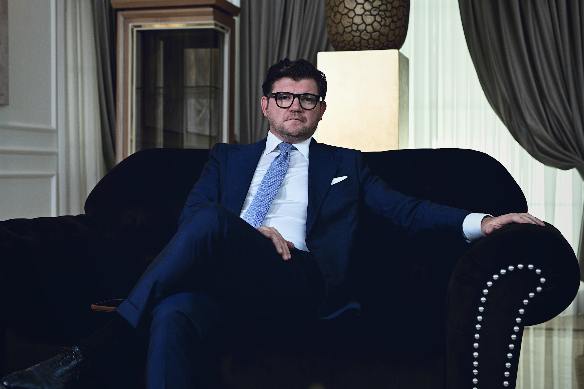Anton Pisaroglu sitting with his legs crossed on a dark sofa in a blue suit and pale blue tie, with one arm along the armrest.