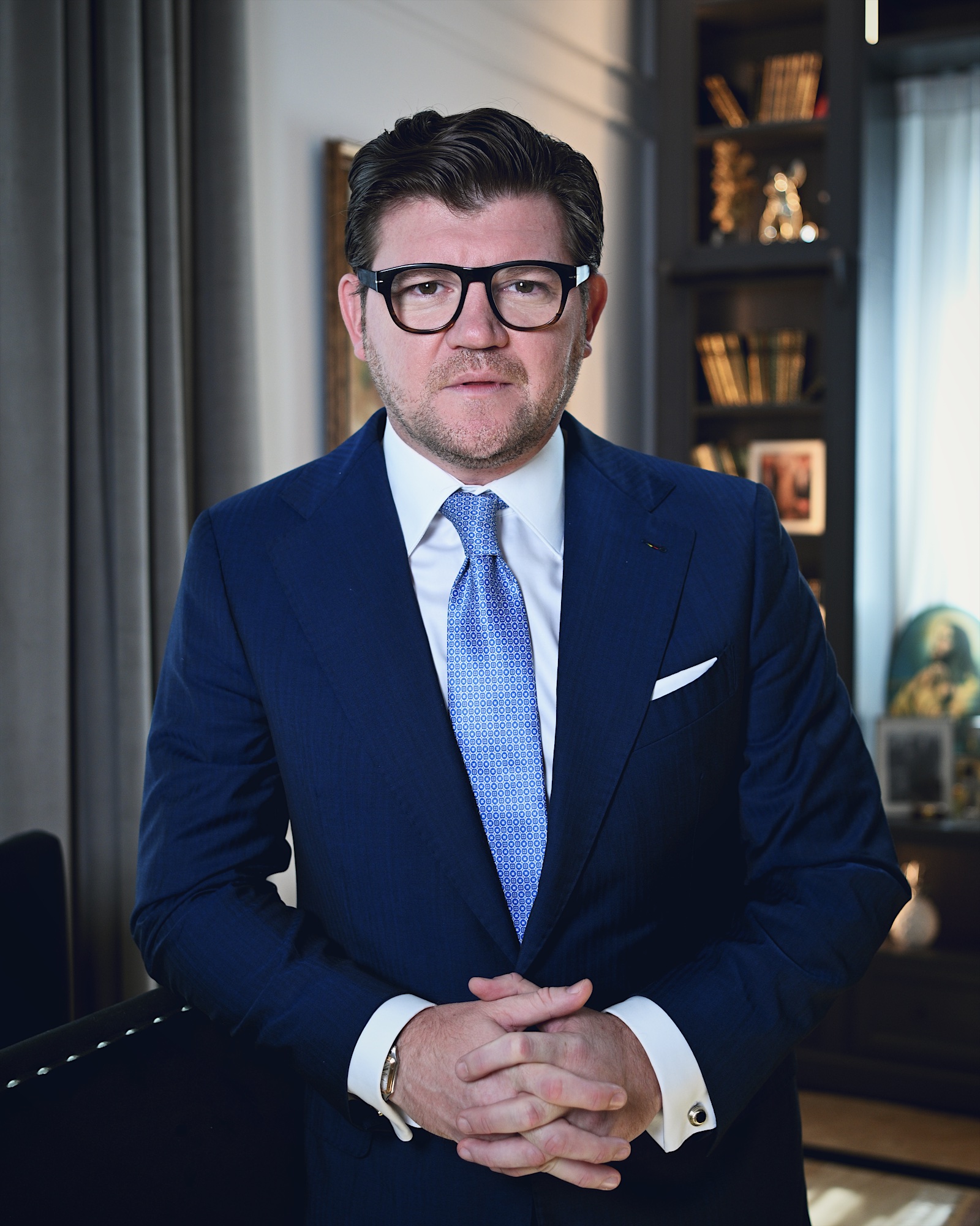 Anton Pisaroglu facing the camera with his hands clasped, wearing glasses, a blue suit and patterned tie, with bookshelves behind him.